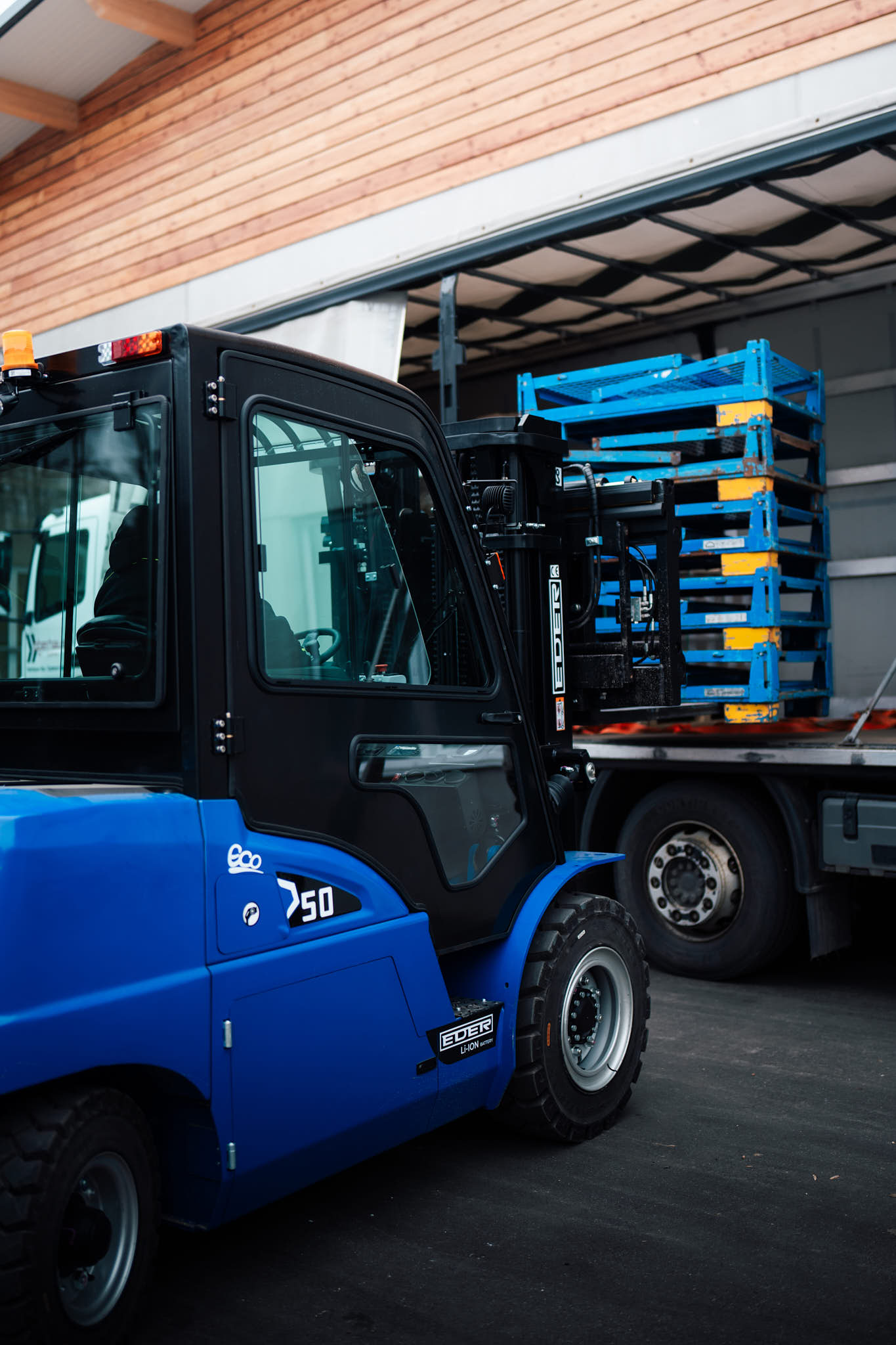 Blauer Eder Eco 50 Li-Ion Elektrostapler beim Entladen gestapelter blauer Paletten von einem Lastwagen vor einer Holzfassade.