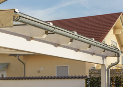 Detailansicht der Entwässerung und Dachkonstruktion eines weißen Holz-Terrassendachs mit Metallrinne bei Tageslicht.