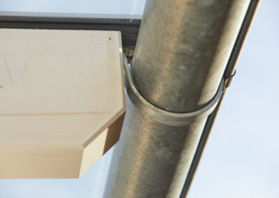 Detailaufnahme eines verzinkten Dachrinnenhalters an einer runden Regenrinne, befestigt an einem Holzsparren eines Terrasse…