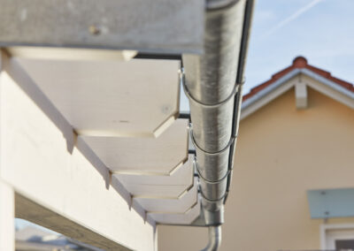 Detaillierte Ansicht einer grauen Regenrinne an einem weißen Holzsparren, Teil einer Terrassenüberdachung an Hausfassade.