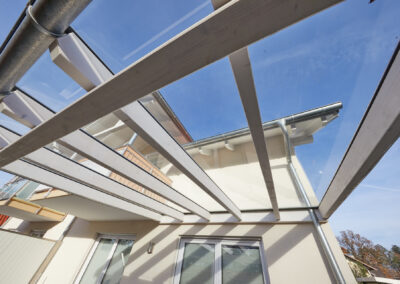 Weiße Holz-Terrassenüberdachung mit Glasdach, direkt an ein helles Gebäude angeschlossen, darunter Fenster und ein Balkon.