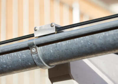 Detailansicht einer verzinkten Regenrinne und einer stabilen Glasklemme, die ein Glaspanel auf einem Terrassendach fixiert.