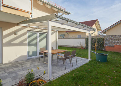 Weiße Holz-Terrassenüberdachung mit transparentem Glasdach über einer Steinterrasse und Gartenmöbeln vor einem modernen Haus.