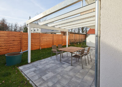 Weiße Holz-Terrassenüberdachung mit Glasdach über gepflasterter Terrasse und Sitzgruppe, inklusive Regenrinne.