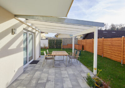 Moderne Terrassenüberdachung aus weißem Holz mit Glasdach und zwei Stützen, integriert an einem Haus.