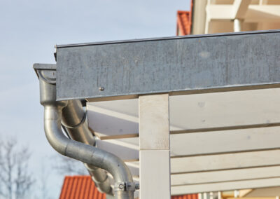 Detailansicht einer verzinkten Regenrinne und Fallrohrs an einem weißen Holz-Terrassendach, effektive Wasserableitung.