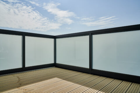 Moderne Balkonbrüstung aus mattem Glas und dunklen Aluminiumprofilen auf Holzdeck unter blauem Himmel.