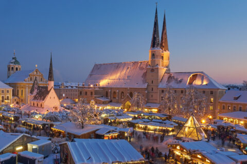 Verschneiter Kapellplatz in Altötting bei Dämmerung mit festlich beleuchtetem Weihnachtsmarkt, Marienbrunnen-Glaspyramide un…