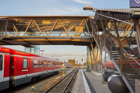Barrierefreie Fußgängerbrücke mit Glaselementen und Treppenhaus über Bahngleisen am Bahnhof Mühldorf, neben einem stehenden…