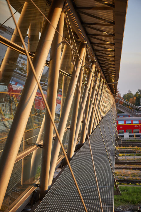 Moderner Bahnübergang in Mühldorf am Inn mit eleganter Glas- und Stahlkonstruktion, die das Sonnenlicht reflektiert.