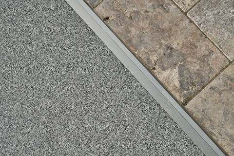 Nahaufnahme des Übergangs von grauer Balkofloor-Paneele aus Aluminium zu beigen Natursteinplatten, getrennt durch ein Metallprofil.