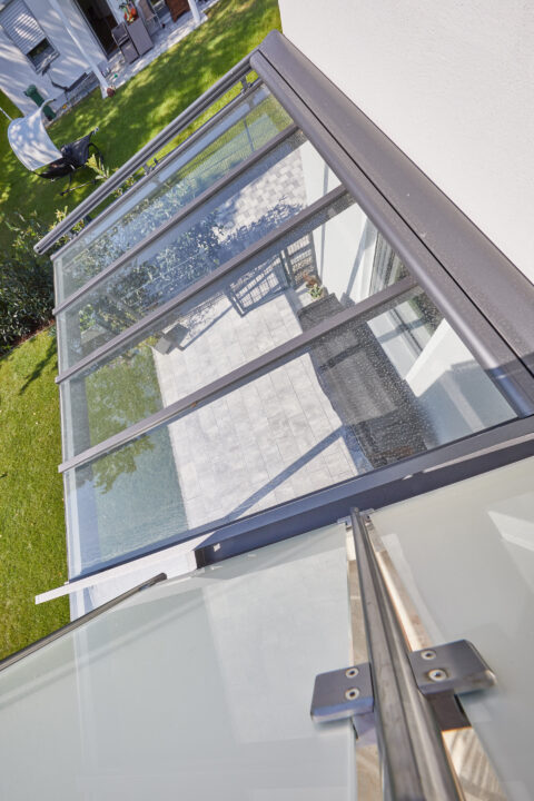 Detaillierte Ansicht des Übergangs eines modernen Glas-Terrassendachs mit dunkler Aluminiumkonstruktion und passender Verble…