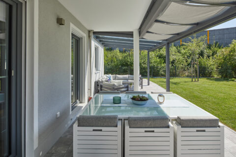 Modernes Glas-Terrassendach über einem eleganten Ess- und Loungebereich im Garten, schützt vor Wetter.