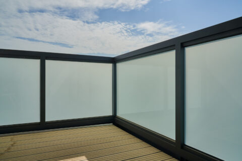 Modernes Balkongeländer aus Aluminium mit mattierten Glasfüllungen auf einer Holzterrasse unter blauem Himmel.