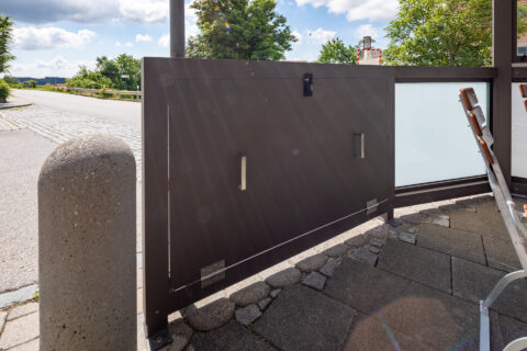 Dunkles Aluminium Balkongeländer mit matter Glasfüllung und integrierter Tür in einem Außenbereich, umgeben von Pflasterstei…