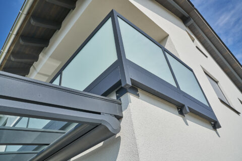 Maßgefertigtes anthrazit pulverbeschichtetes Balkongeländer aus Glas und moderne Terrassenüberdachung an einem weißen Gebäud…