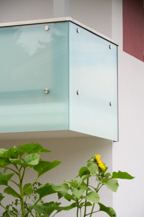 Mattglas-Balkonverglasung mit Edelstahl-Punkthaltern an weißer Fassade.