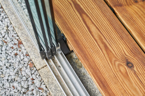Detailansicht der Befestigung einer Bodenschiene für eine Glasschiebeanlage auf einer Holzterrasse mit Granitpflaster und Ki…