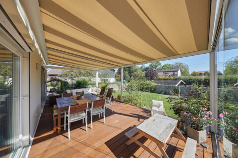 Weinor Sottezza Unterdachmarkise beschattet modernen Kaltwintergarten mit Holzterrasse, Essbereich und Blick in den Garten.