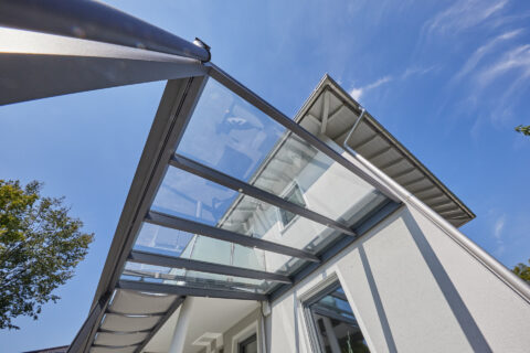 Blick von unten auf eine moderne Terrassenüberdachung aus Glas und Metall, montiert an einem Einfamilienhaus.