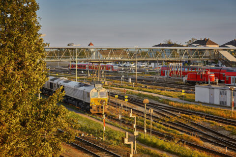 Moderne Fußgängerbrücke aus Glas und Stahl überspannt mehrere Gleisanlagen am Bahnhof Mühldorf am Inn mit Zügen.