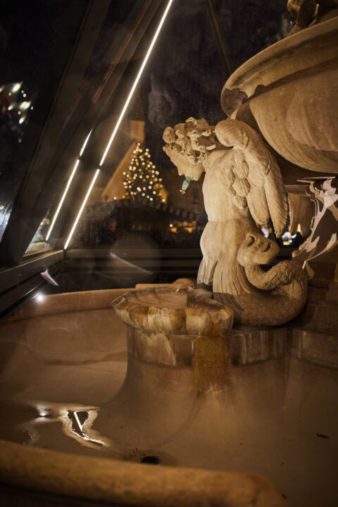 Brunneneinhausung aus Glas mit LED-Beleuchtung schützt eine historische Brunnenfigur mit Engel und Fischschwanz in nächtlich…