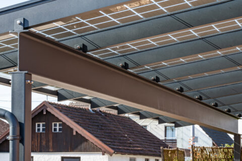 Nahaufnahme eines modernen Photovoltaik-Carports mit robusten T-Trägern und Solarpanels über einem Einfamilienhaus.