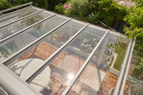 Modernes filigranes Terrassendach aus Klarglas über einer Holzterrasse mit Lounge-Möbeln und Blumenbeeten im grünen Garten.