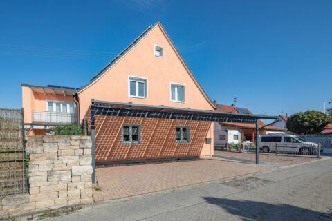 Moderner Photovoltaik Glasdach Carport vor orangefarbenem Einfamilienhaus, erzeugt saubere Energie und bietet Wetterschutz.