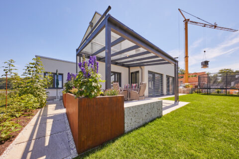 Eckansicht einer modernen Aluminium-Terrassenüberdachung mit Glasdach und filigraner Dachrinne an einem Einfamilienhaus.