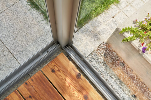 Detailansicht des Eckpfostens und der Bodenschiene einer Kaltwintergarten-Schiebeanlage aus Glas, Holzterrasse und Pflaster.