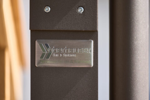Metallplakette 'Oberhauser Bau Systeme' an einer braunen Terrassenüberdachung aus eigener Herstellung, Projekt Niederbergkir…
