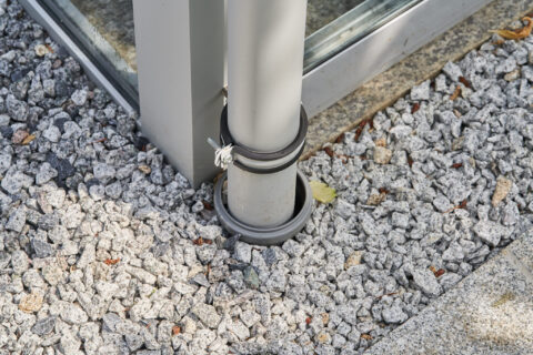 Detailaufnahme der Regenwasserentwässerung eines Aluminium-Kaltwintergartens in Zorneding mit Kiesbett und stabiler Rohrbefe…