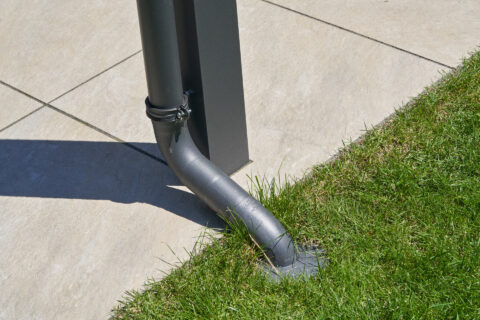 Fallrohr mit Bogen zur Entwässerung einer modernen Terrassenüberdachung, präzise in Rasen neben Terrassenplatten.