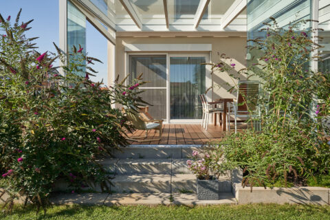 Moderne, filigrane Glas-Aluminium Terrassenüberdachung mit Holzdeck und gemütlicher Sitzecke, umgeben von blühenden Sträuche…