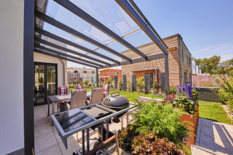 Moderne Terrassenüberdachung aus Glas und Aluminium an einem Haus mit Holzfassade, darunter Esstisch und Grill.