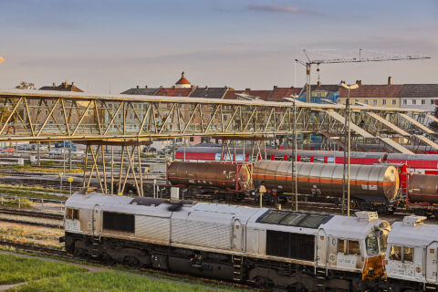 Moderne Fußgängerbrücke mit gläsernem Tunnel über belebten Gleisanlagen des Bahnhofs Mühldorf, verbindet Stadtteile sicher.