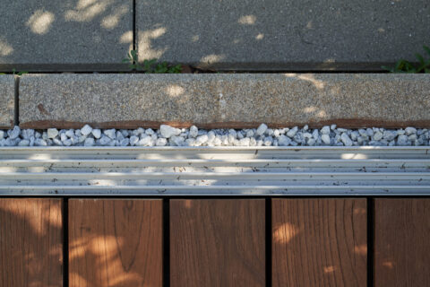 Nahaufnahme einer versenkten, barrierefreien Bodenschiene für eine Ganzglasschiebeanlage zwischen Holzterrasse und Pflasters…