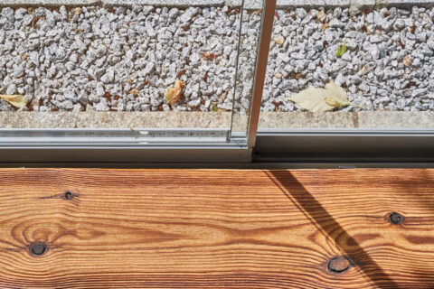 Detaillierte Ansicht einer barrierefreien, versenkten Ganzglas-Schiebeanlage mit Holzterrasse und Kiesbett für nahtlose Über…