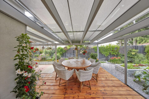 Moderner Glaswintergarten mit Holzterrasse, runden Esstisch und Korbstühlen, darunter ein flexibles Beschattungssystem. Blic…