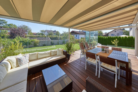 Moderner Kaltwintergarten mit Glasdach und Markise auf Holzterrasse, komfortable Outdoor-Möbel und Gartenblick in Oberpframm…