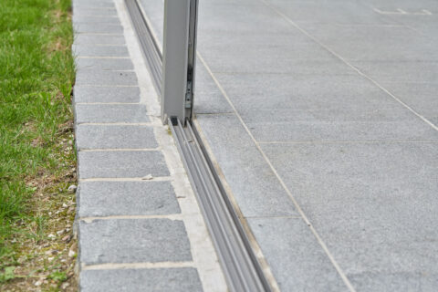Detaillierte Ansicht der Bodenschiene einer Glasschiebetür für Terrassenüberdachung, angrenzend an Pflastersteine und Rasen.