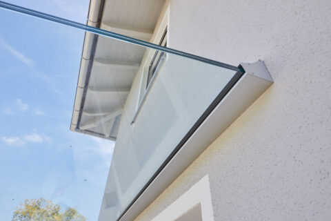 Moderne Glas-Eingangsüberdachung an einer hellen Hauswand, klares Design, Schutz vor Witterungseinflüssen, elegante Ästhetik.