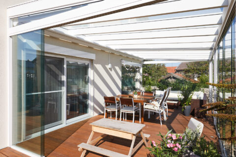 Moderne Glas-Schiebewand für eine Terrassenüberdachung mit weißem Rahmen, Holzboden, Esstisch und Lounge-Möbeln in Oberpfram…