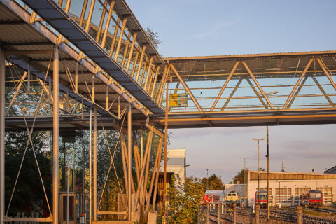 Glas-Stahl-Fußgängerbrücke am Bahnhof Mühldorf über Bahngleisen im Sonnenlicht, mit Zügen und Gebäuden im Hintergrund.