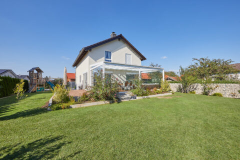 Modernes Einfamilienhaus mit Glas-Wintergarten und integriertem Sonnenschutz in Oberpframmern, umgeben von einem gepflegten…