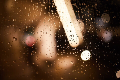 Regentropfen auf einer Glasscheibe mit spiegelnden Lichtern im Hintergrund.