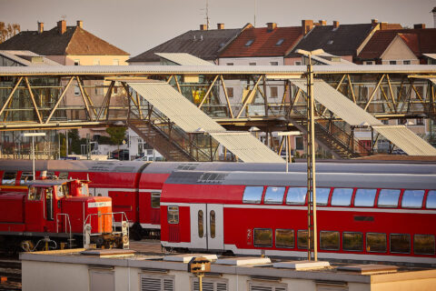 Architektonischer Glasbau mit Treppenaufgang und Überführung an einem modernen Bahnhof, unter dem rote Züge parken. Zeigt Ob…