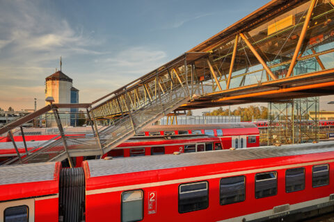 Fußgänger-Glasbrücke aus Stahl und Glas über Bahngleise mit roten Zügen am Bahnhof Mühldorf, mit Treppenzugang und Wolken am…
