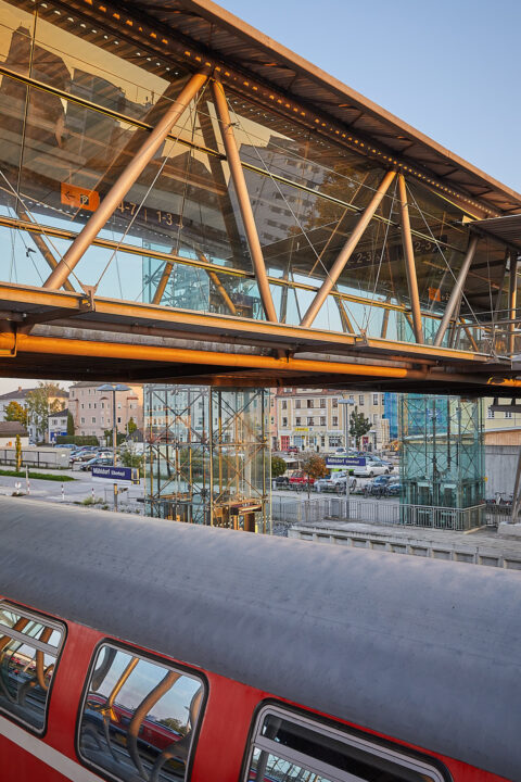 Moderne Glas-Stahl-Fußgängerbrücke über Gleise am Bahnhof Mühldorf bei Sonnenlicht, mit Aufzügen und Zug.